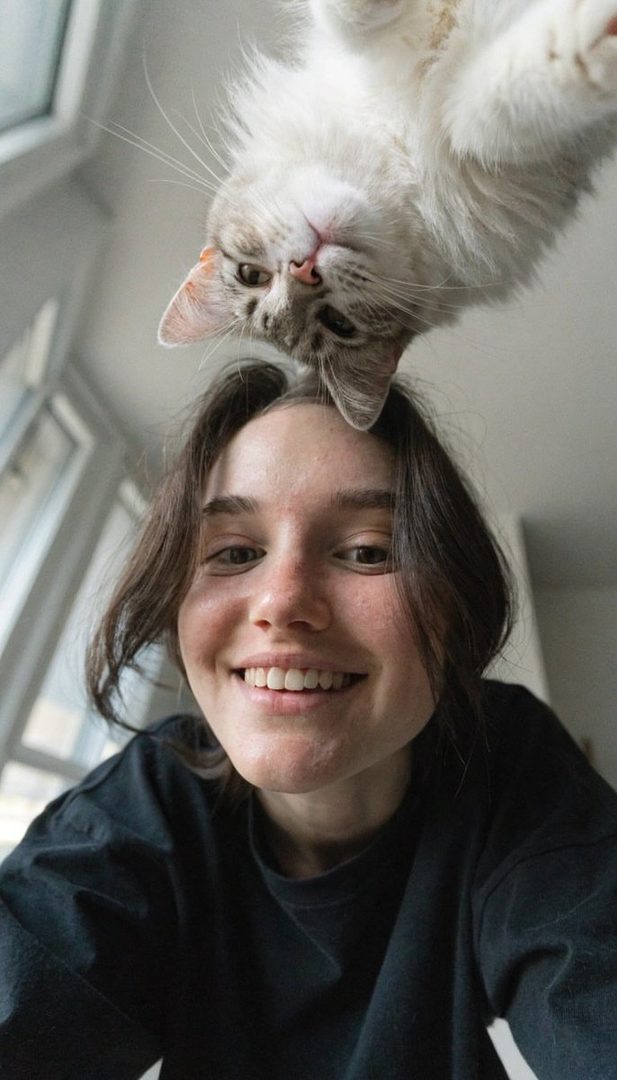 A young woman with a warm smile and a fluffy cat perched on her head, taken from a low angle in a bright indoor setting.