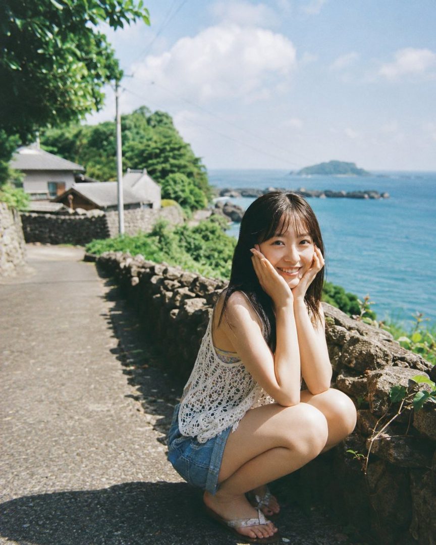 A young Japanese woman with long black hair smiles while crouching on a stone path overlooking the ocean.