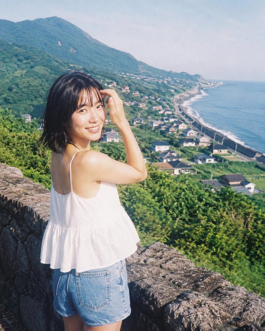 A young woman with short black hair smiles at the camera, standing on a stone wall overlooking a coastal town and the ocean.
