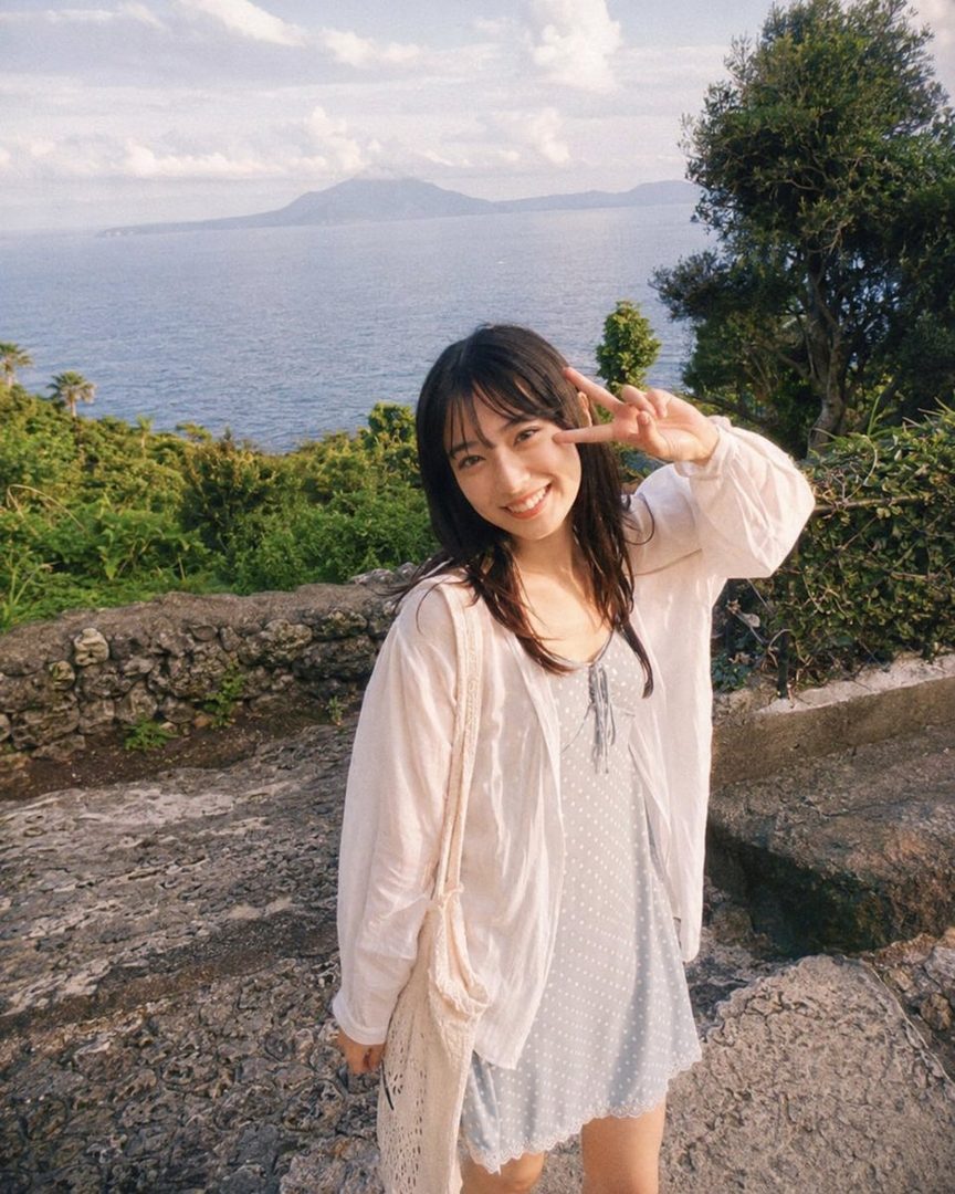A young Japanese woman with long black hair smiles and makes a peace sign at the camera, wearing a light blue polka dot dress and a white open cardigan, with a scenic ocean and mountain view in the background.