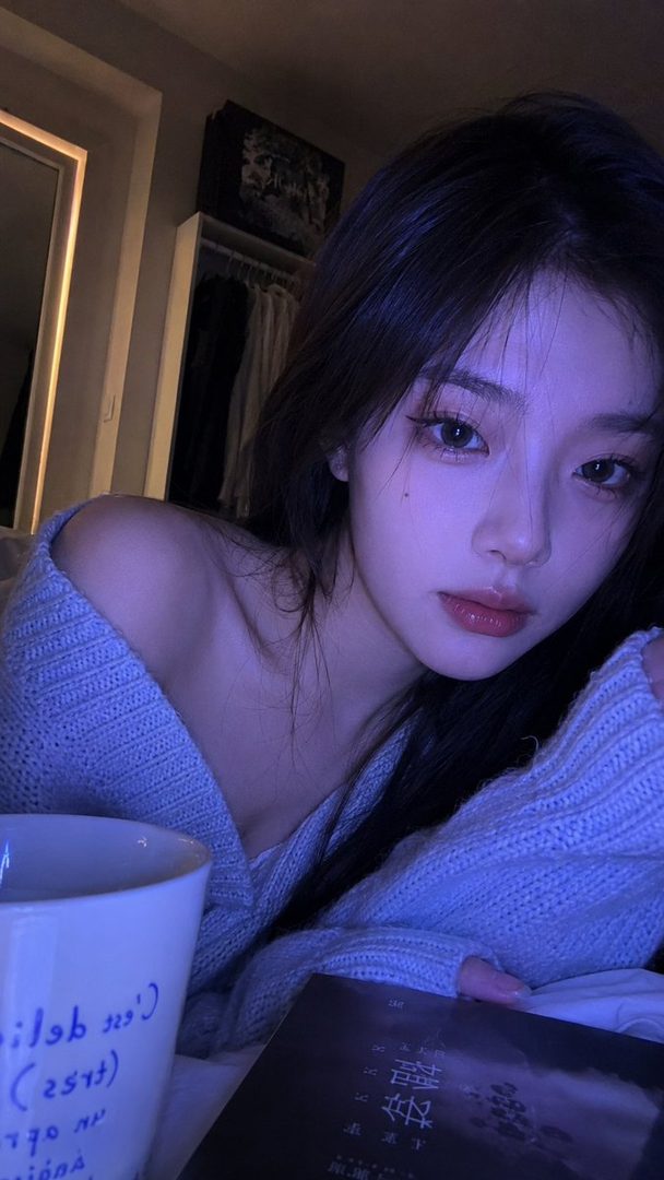 A young East Asian woman with long dark hair, wearing a light blue off-the-shoulder sweater, poses for a selfie indoors with a book and a mug.