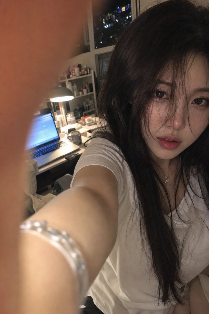 A young woman takes a close-up selfie in her room at night, with her arm and finger partially obscuring the lens, a laptop and cluttered desk in the background.