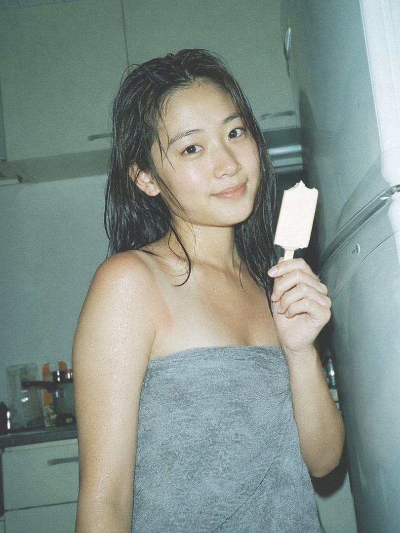 A young woman with wet hair, wrapped in a towel, holds a bitten popsicle and smiles.