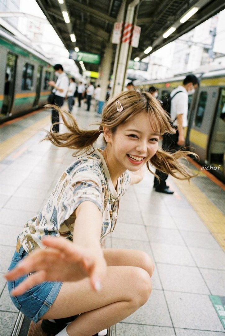 A young woman with blonde twintails smiles brightly while crouching on a train platform.