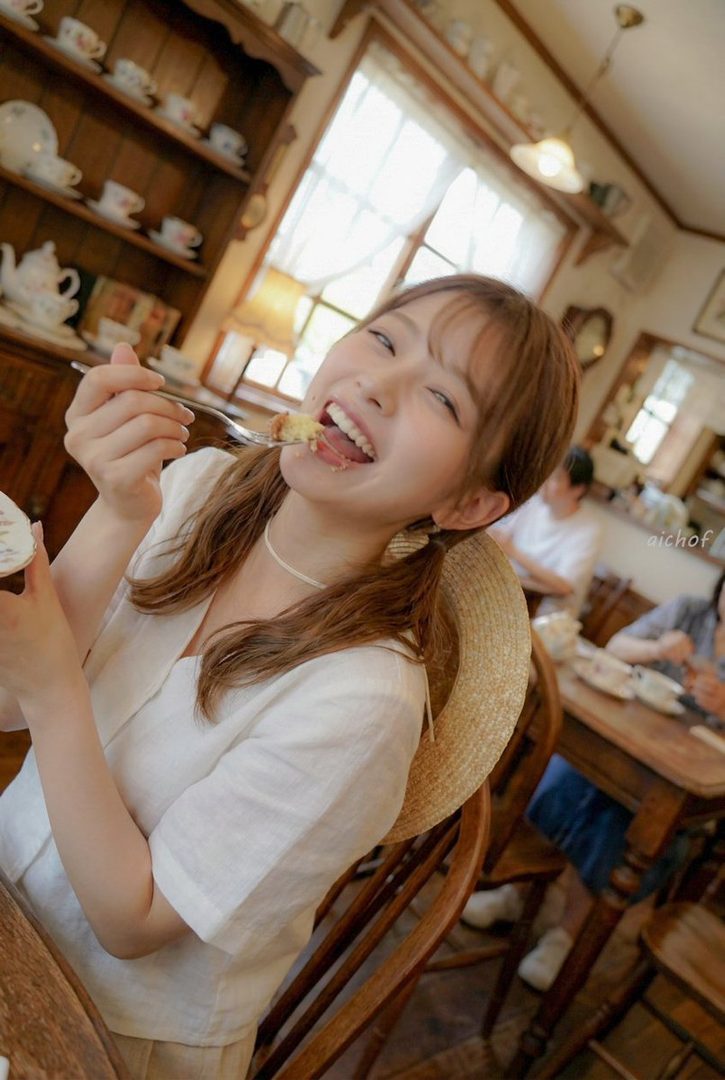 A young woman with brown hair in twintails smiles widely while eating cake at a cafe.