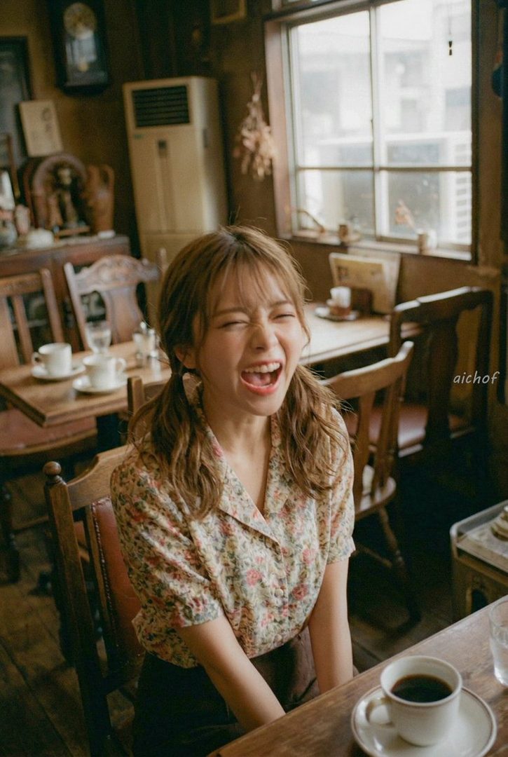 A young woman with brown hair in pigtails, wearing a floral shirt, laughs heartily while sitting at a table in a cafe.