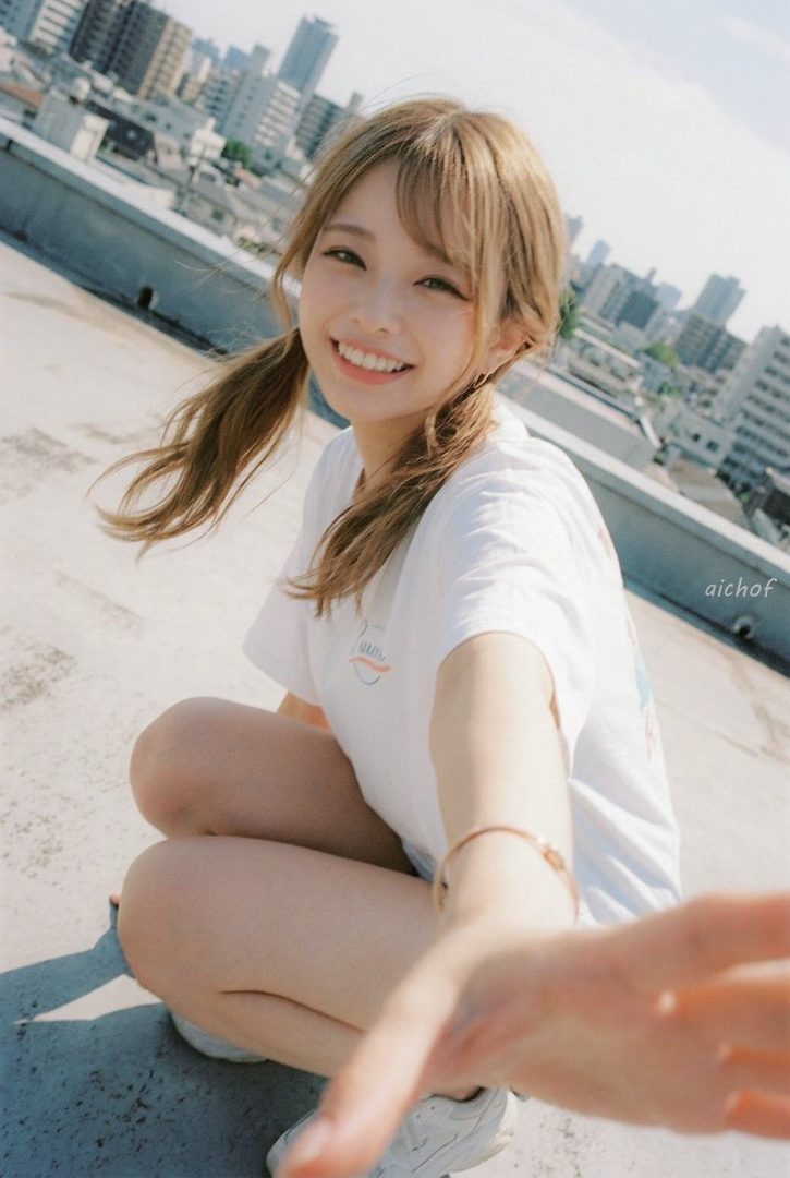 A young woman with blonde twin-tails smiles at the camera while crouching on a rooftop with a city skyline in the background.