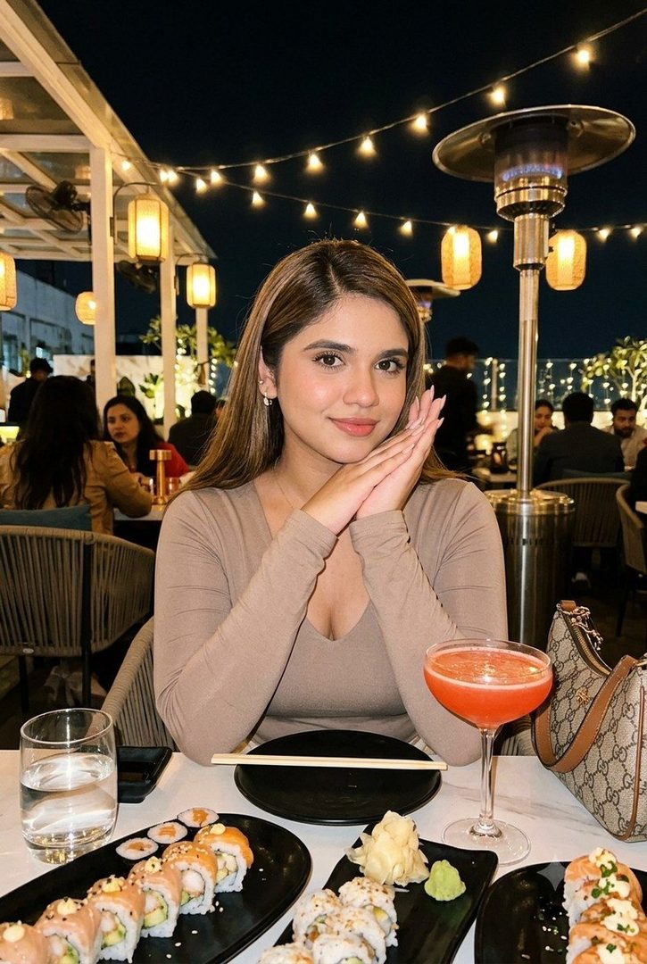 A young woman with long brown hair smiles gently at the camera while sitting at a restaurant table with sushi and a cocktail.
