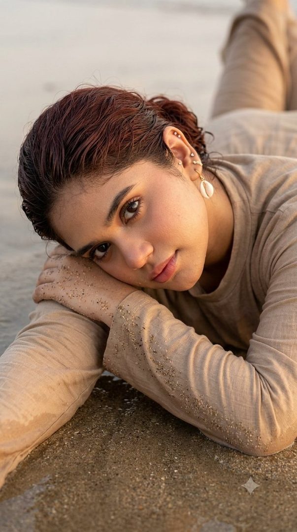 A close-up portrait of a young woman lying on a sandy beach, resting her head on her arms, with sand particles on her skin and clothing.