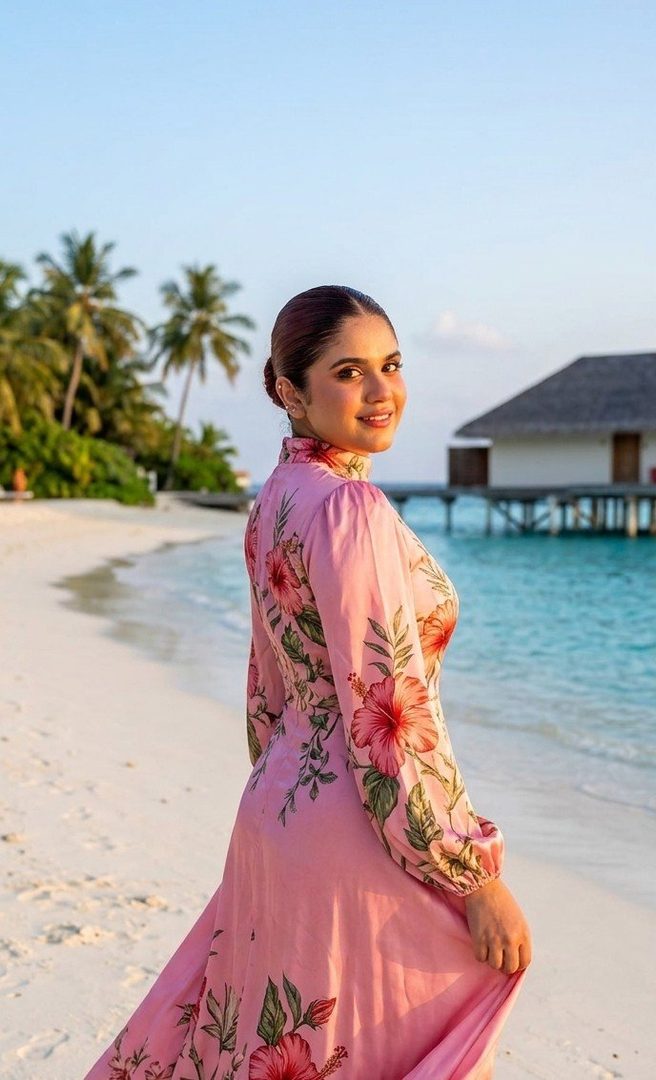 A woman in a pink floral dress stands on a beach at sunset, looking over her shoulder.