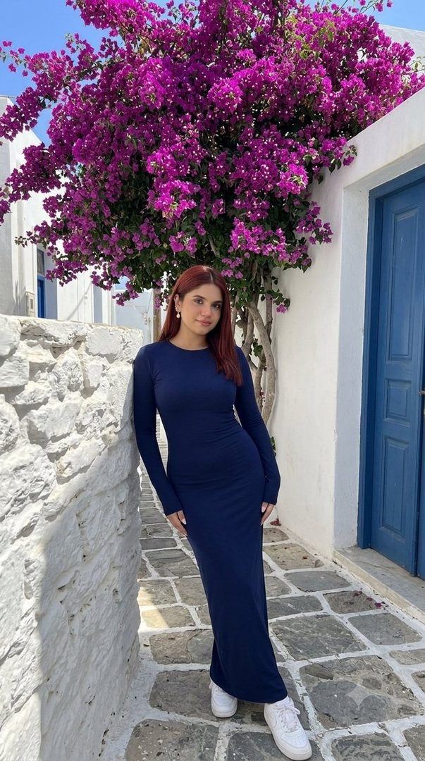 A young woman with long brown hair, wearing a navy blue long-sleeved dress and white sneakers, stands in a narrow Greek street with a whitewashed stone wall and a vibrant bougainvillea tree.