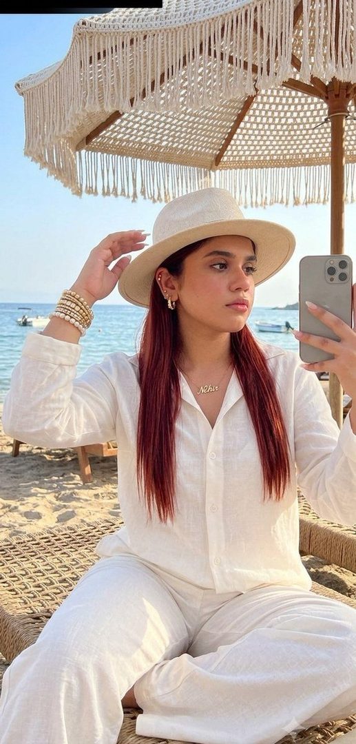 A woman with long red hair and a hat takes a selfie on a beach, wearing a white shirt and pants, with the sea and a bohemian umbrella in the background.