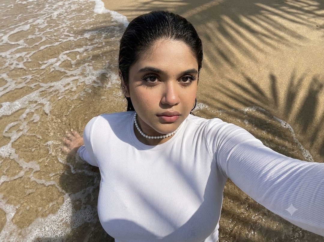 A young woman with dark hair, wearing a white long-sleeved top and a pearl choker, takes a selfie while lying on the surf line at a sandy beach, with palm tree shadows dappling the scene.