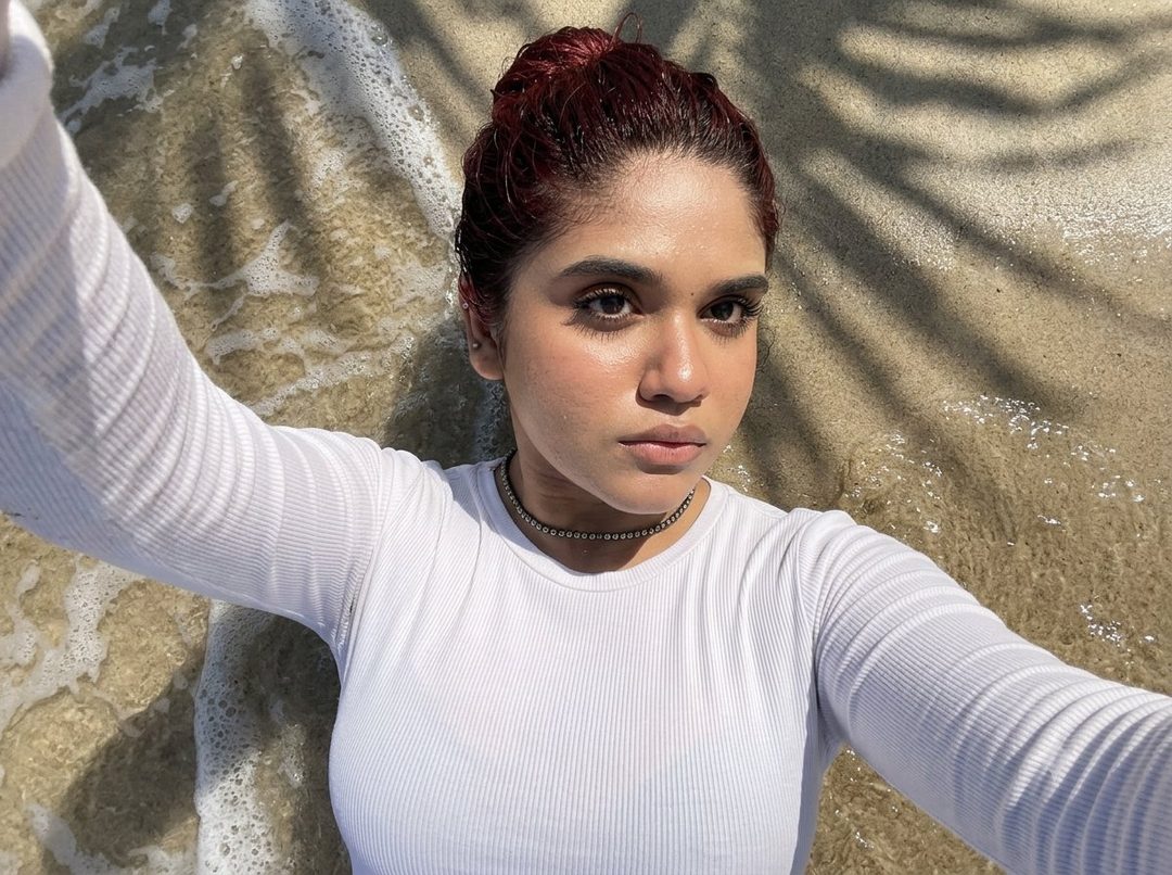 A young woman with dark red hair tied up in a bun, wearing a white long-sleeved top and a choker necklace, takes a selfie while lying on the surf line at the beach.