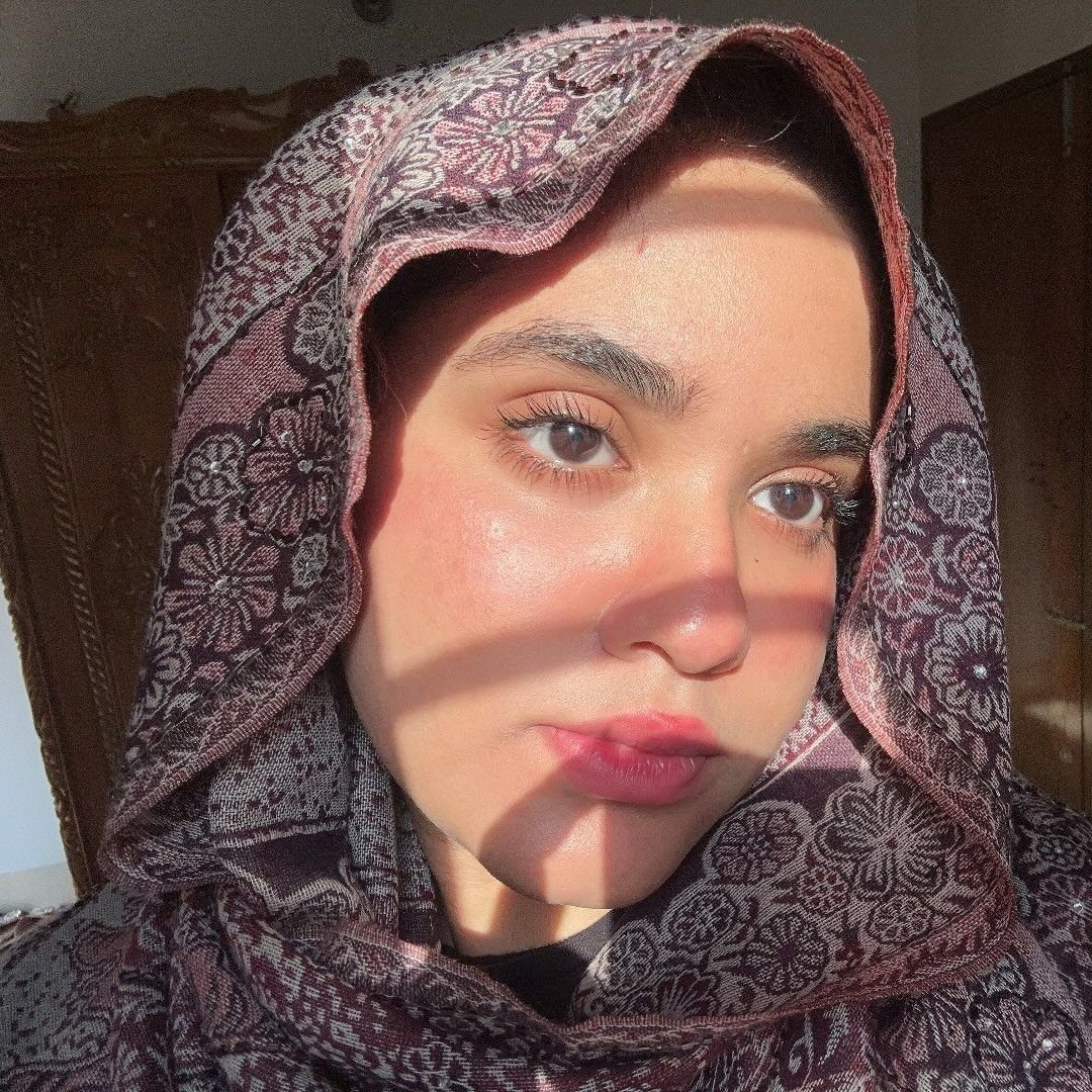 A young woman wearing a patterned headscarf poses for a selfie, with sunlight casting shadows across her face.