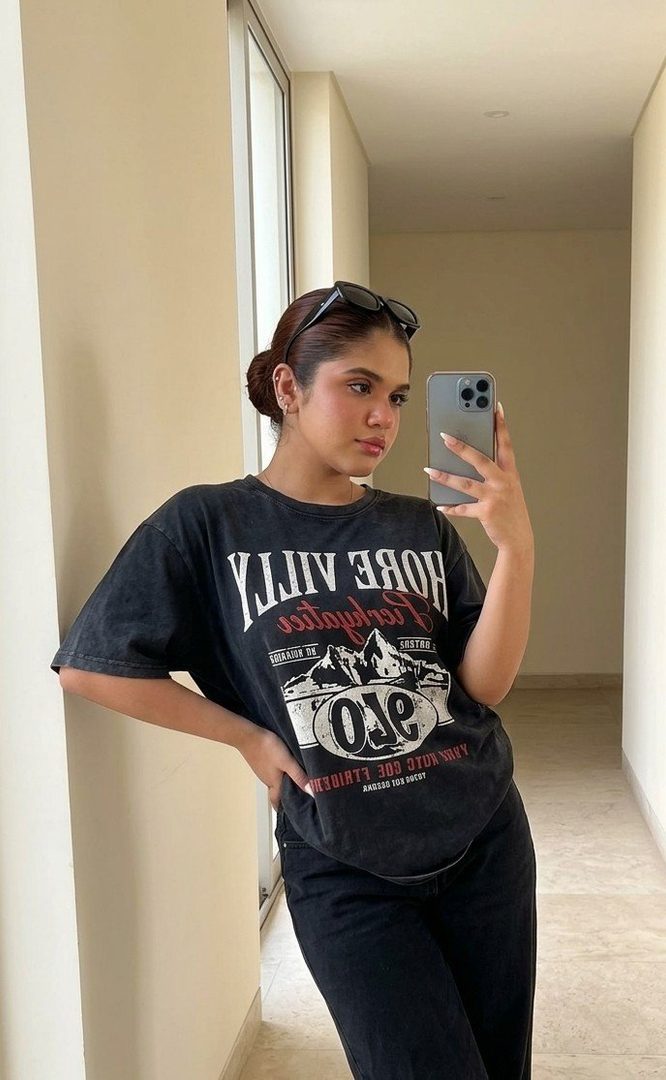A young woman takes a mirror selfie in a hallway, wearing an oversized graphic t-shirt and baggy jeans, with sunglasses perched on her head.