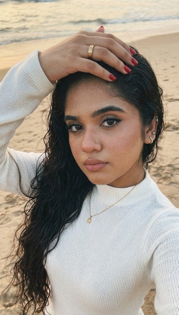 A young woman with long, wavy black hair and a white knitted sweater takes a selfie on a sandy beach at sunset, looking into the camera with a relaxed expression.