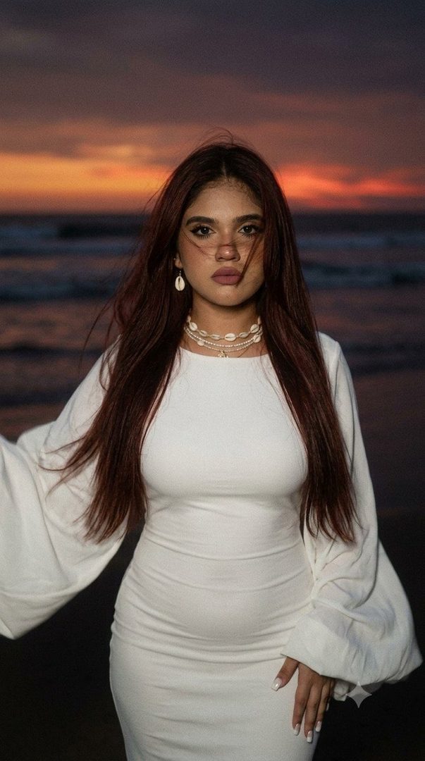 A woman in a white dress stands on the beach at sunset, with her long hair blowing in the wind.