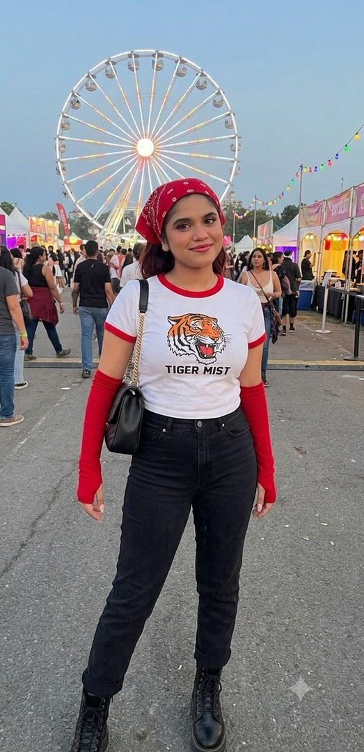 A young woman in a white tiger print t-shirt and red bandana smiles at the camera at a lively outdoor festival with a Ferris wheel in the background.