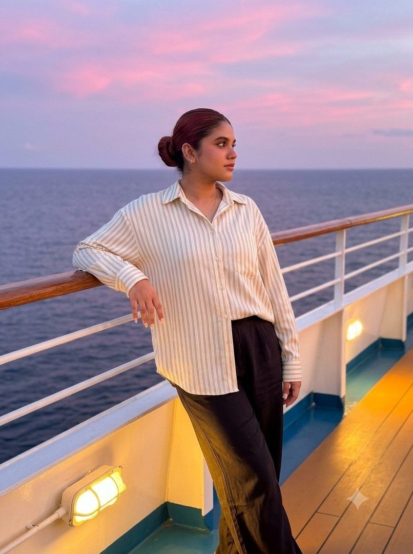 A young woman with her hair in a bun stands on a cruise ship deck at sunset, leaning on the railing and looking out at the ocean.