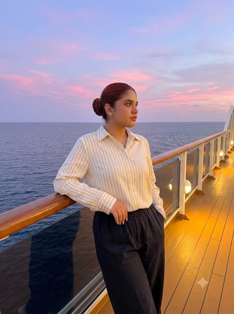 A young woman with her hair in a bun stands on a cruise ship deck at sunset, wearing a striped shirt and dark trousers, looking out at the ocean.