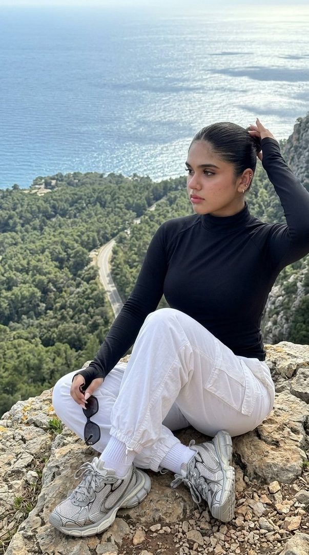 A young woman with dark hair pulled back sits on a rocky cliff overlooking the sea, wearing a black turtleneck and white joggers, with a scenic mountain and ocean view.