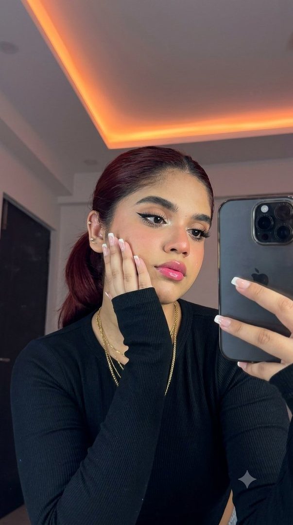 A young woman with dark hair pulled back in a ponytail takes a selfie, resting her cheek on her hand. She wears a black top and gold necklaces, with dramatic eye makeup and glossy pink lips. The background features a ceiling with orange neon lighting.