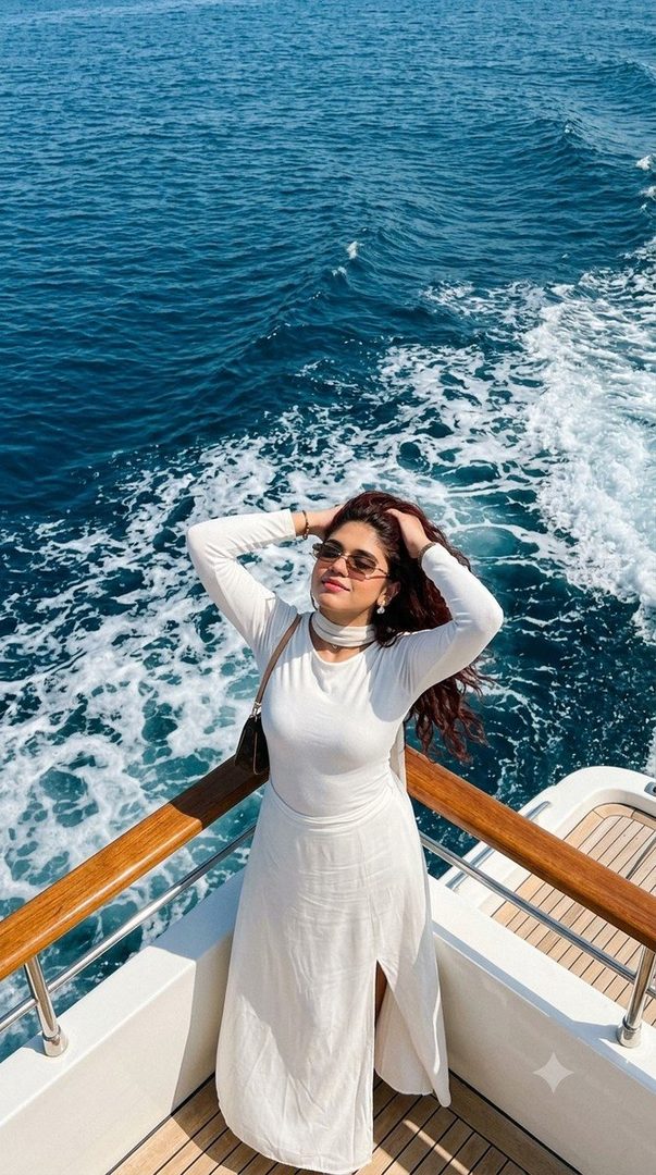 A woman in a white dress stands on a yacht, with her hands in her hair, against the backdrop of the blue sea and white foam.