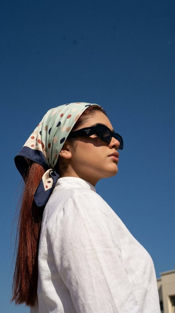 A woman with long brown hair, wearing a white blouse and sunglasses, with a patterned headscarf, looking to the side against a clear blue sky.
