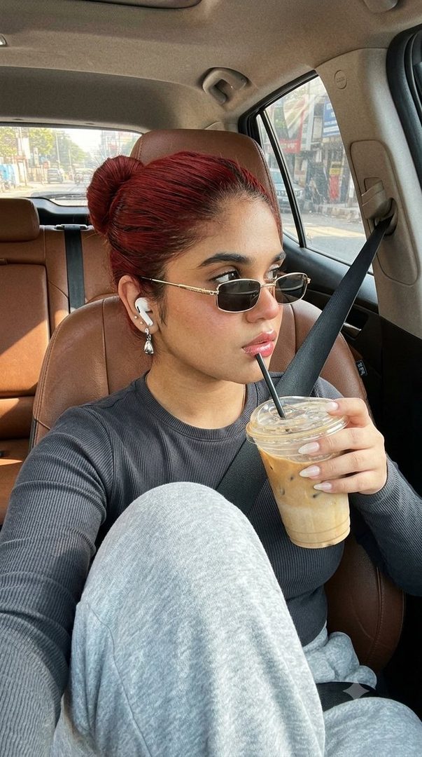 A young woman with red hair in a bun, wearing sunglasses and headphones, drinks a latte while sitting in a car with a blurred urban background.