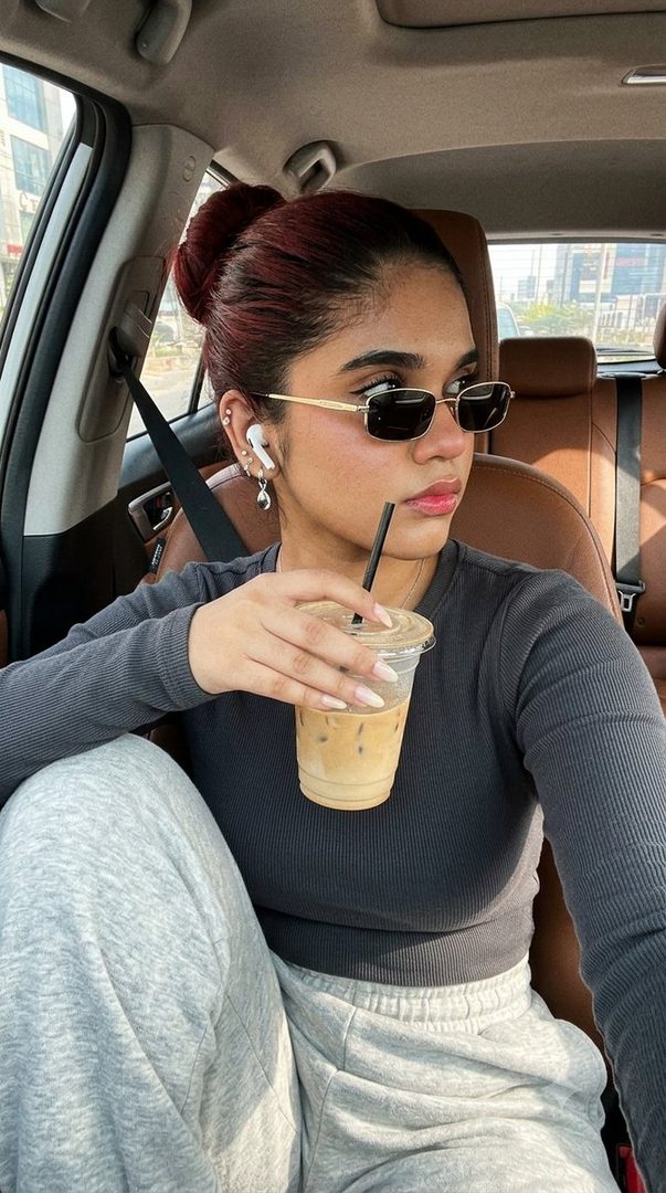 A young woman with her hair in a bun, wearing sunglasses and casual clothes, sits in the driver's seat of a car holding a latte.