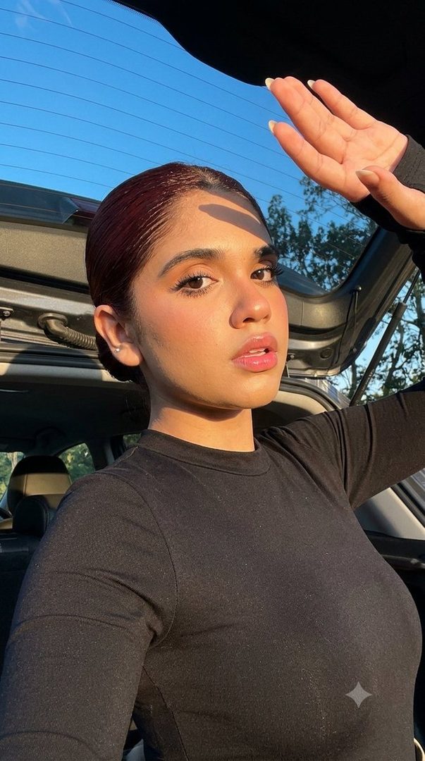 A young woman with brown hair pulled back, wearing a black long-sleeved shirt, poses for a selfie in a car with the hatch open, shielding her face from the sun with her hand.