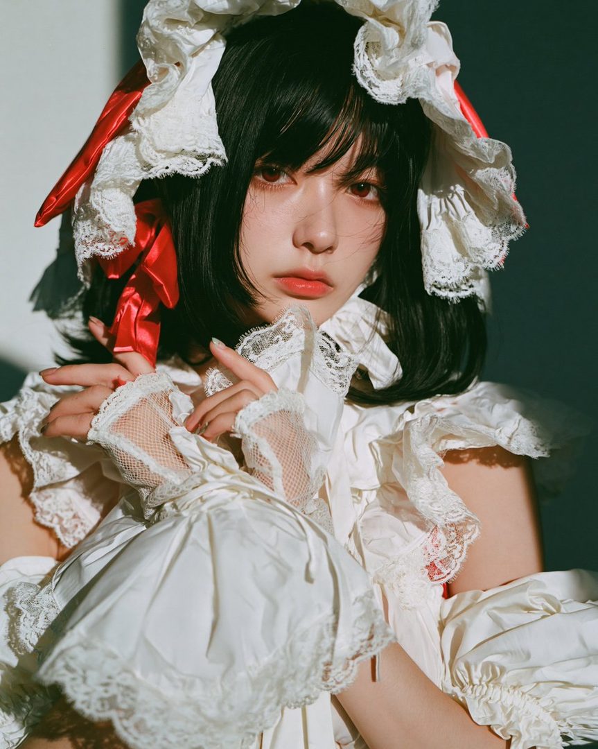 A young woman with black hair and striking red eyes, dressed in an elaborate white and red Victorian-style outfit with lace and ribbons, posing indoors with dramatic lighting.