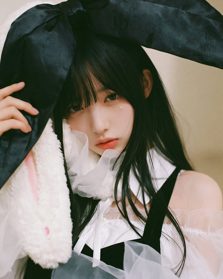 A young woman with long black hair and bangs, wearing a white blouse and black suspenders, with a large black bow and plush bunny ears, posing indoors.