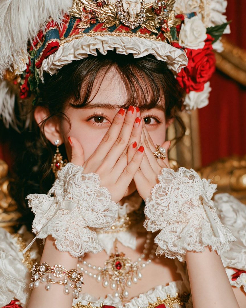 A young woman in elaborate historical costume and headdress, covering her mouth with her hands.