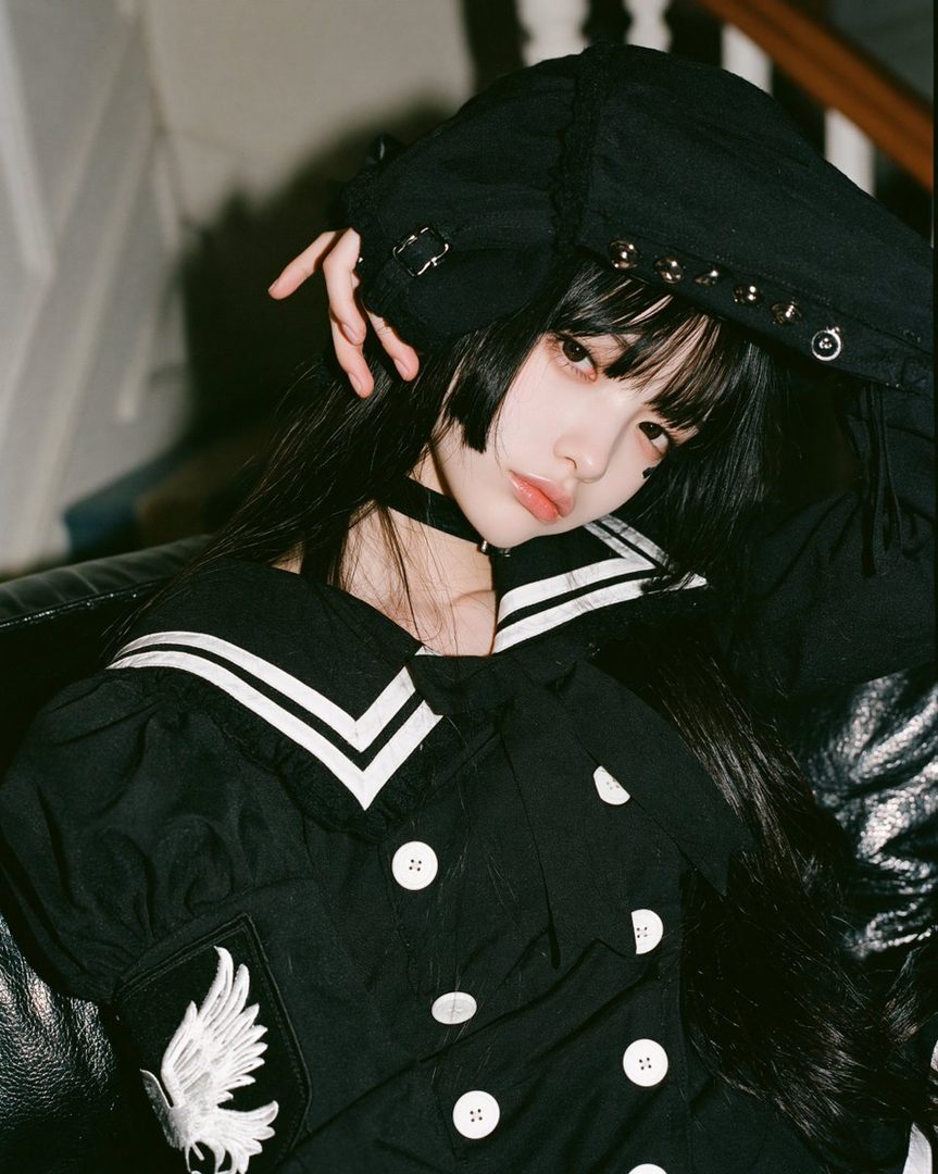 A young woman with long black hair and bangs, wearing a black sailor-style outfit and a choker, poses indoors.