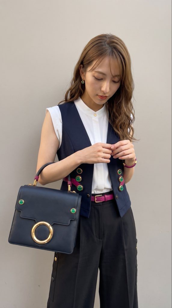A young East Asian woman with long brown hair, wearing a white blouse, a navy vest, and black pants, holding a dark handbag.