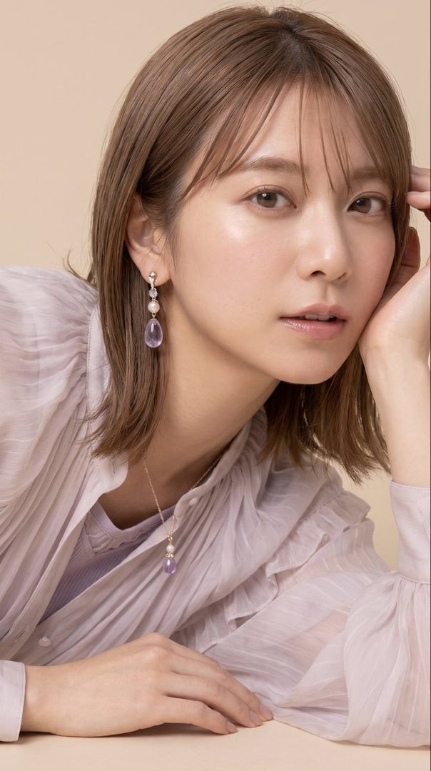 A young East Asian woman with short brown hair and bangs, wearing a light purple blouse and delicate jewelry, poses elegantly indoors.