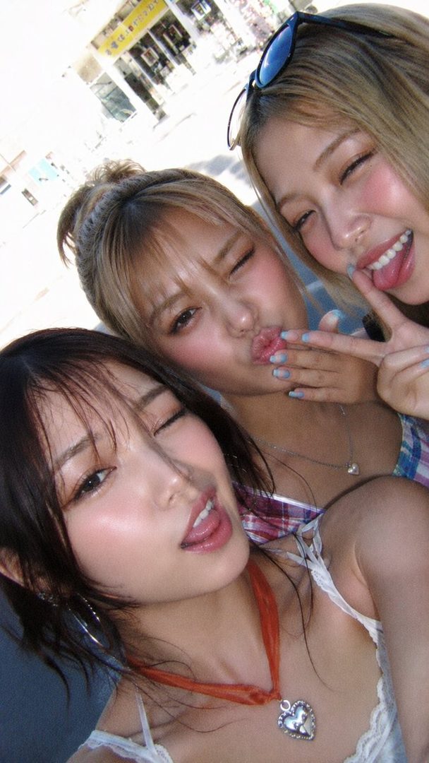 Three young East Asian women taking a selfie outdoors, making playful expressions.