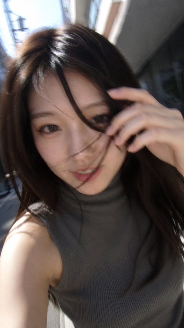 A young woman with long brown hair touches her hair while looking at the camera, outdoors.