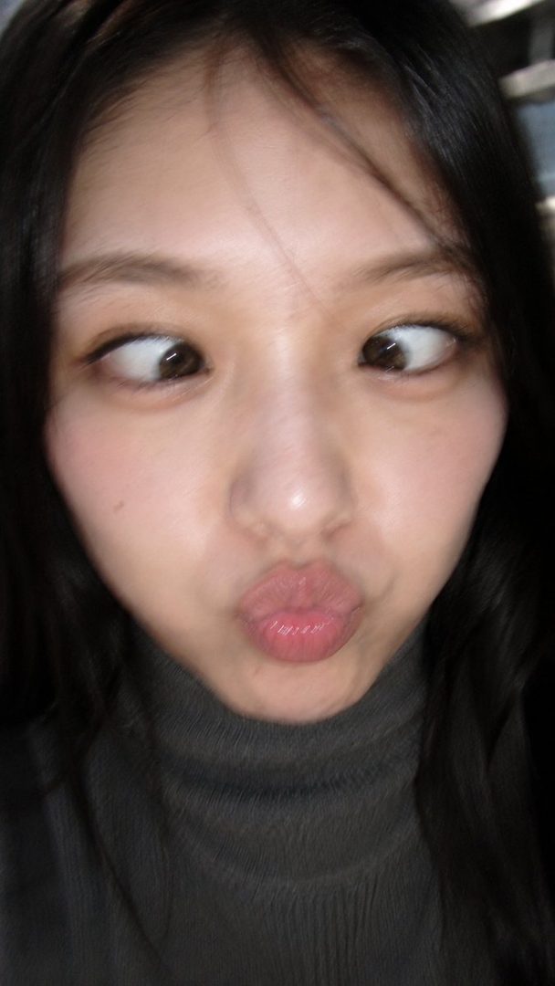 A young East Asian woman with dark hair makes a funny face, crossing her eyes and puckering her lips, while wearing a dark turtleneck sweater.