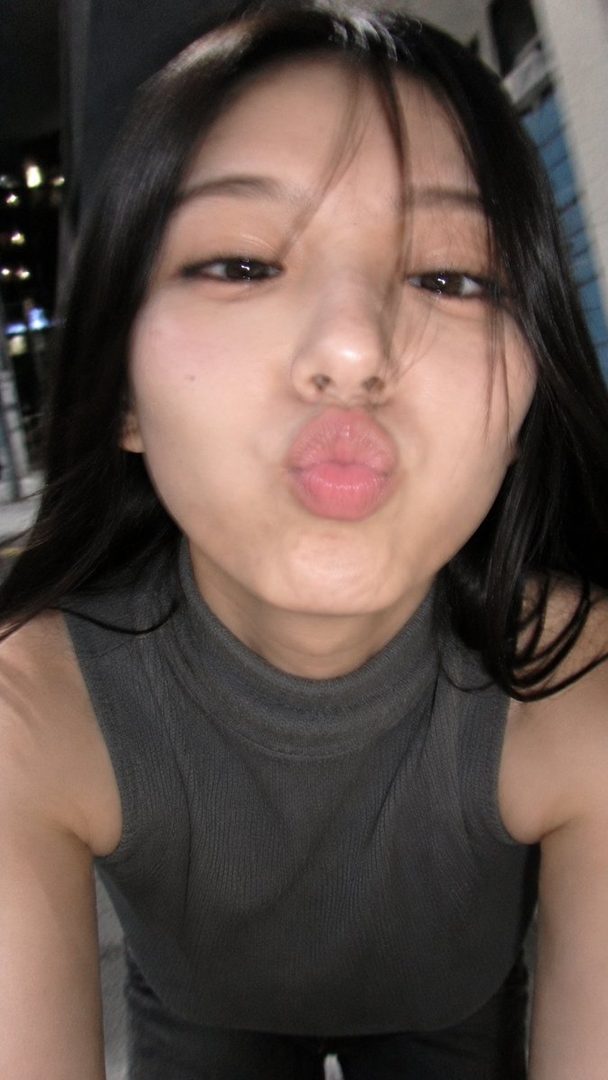 A young East Asian woman with long black hair makes a kissing face towards the camera in a close-up selfie taken outdoors at night.