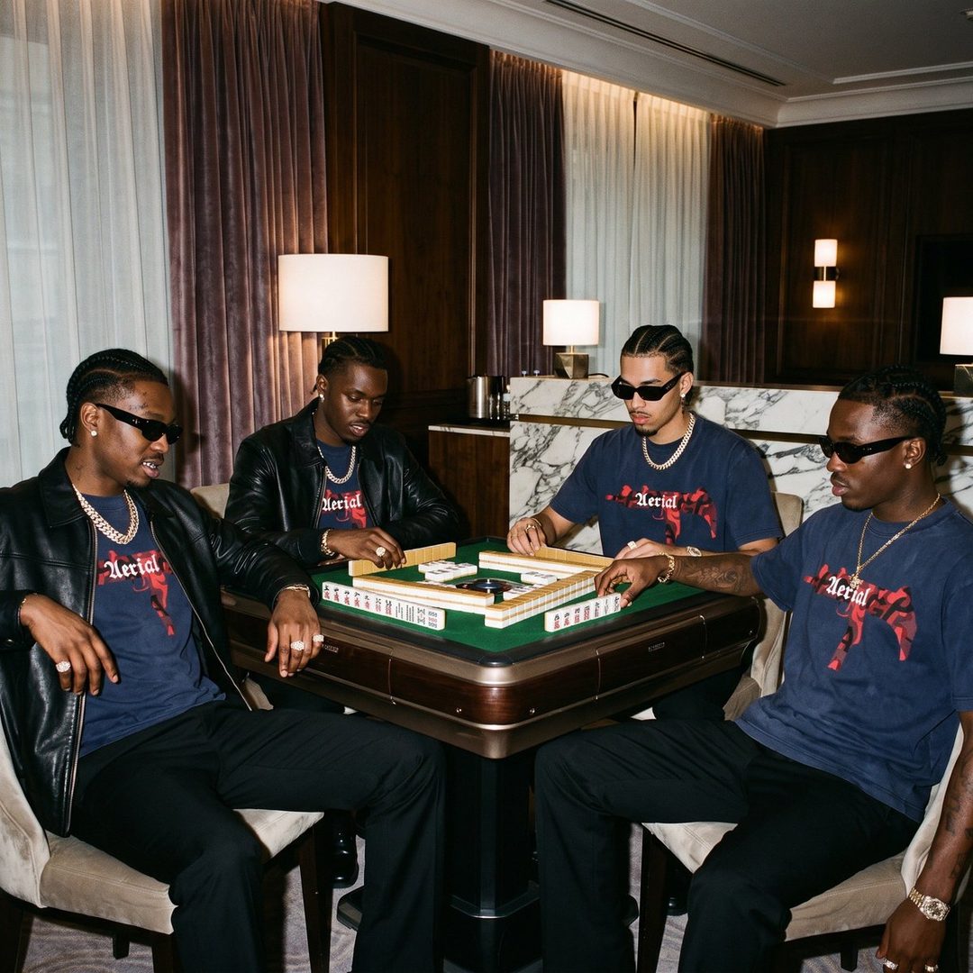 Four young men playing mahjong in a luxurious indoor setting, wearing matching graphic t-shirts and sunglasses.