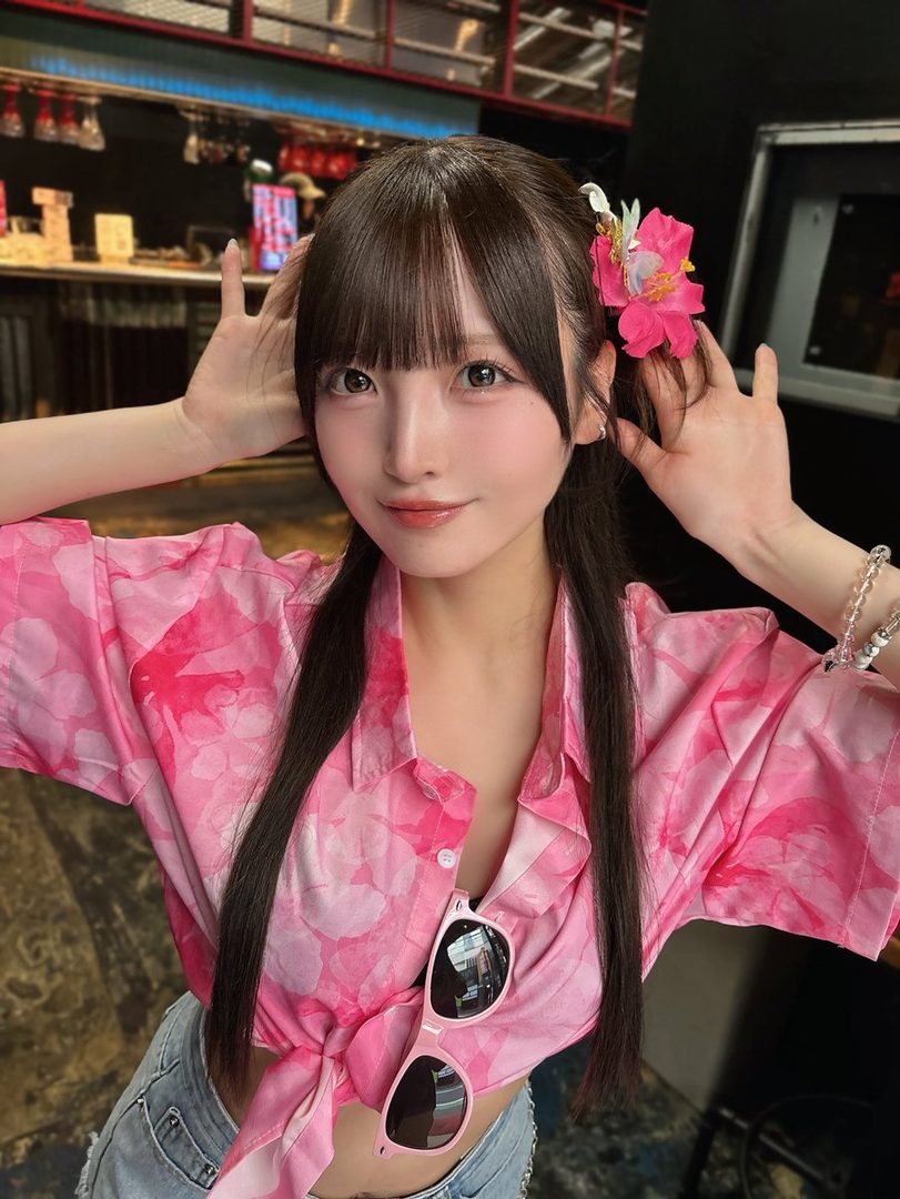 A young East Asian woman with long dark hair and bangs, wearing a pink floral print shirt tied at the waist, poses for a selfie with a flower in her hair.
