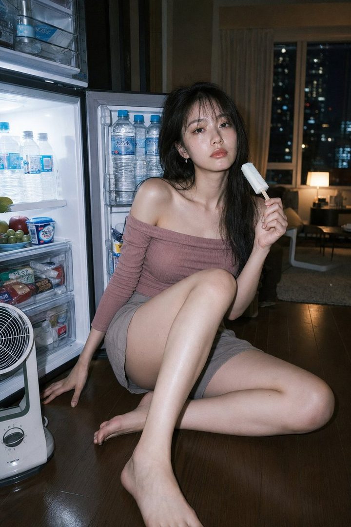 A young East Asian woman with long dark hair sits on the floor next to an open refrigerator, holding an ice pop, in a dimly lit apartment at night.