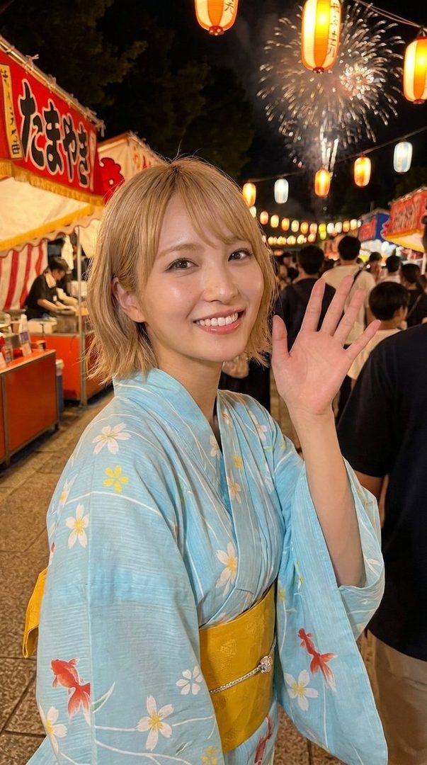 A young Japanese woman with short blonde hair smiles and waves, wearing a light blue yukata with floral and goldfish patterns, at a summer festival at night with lanterns and fireworks.