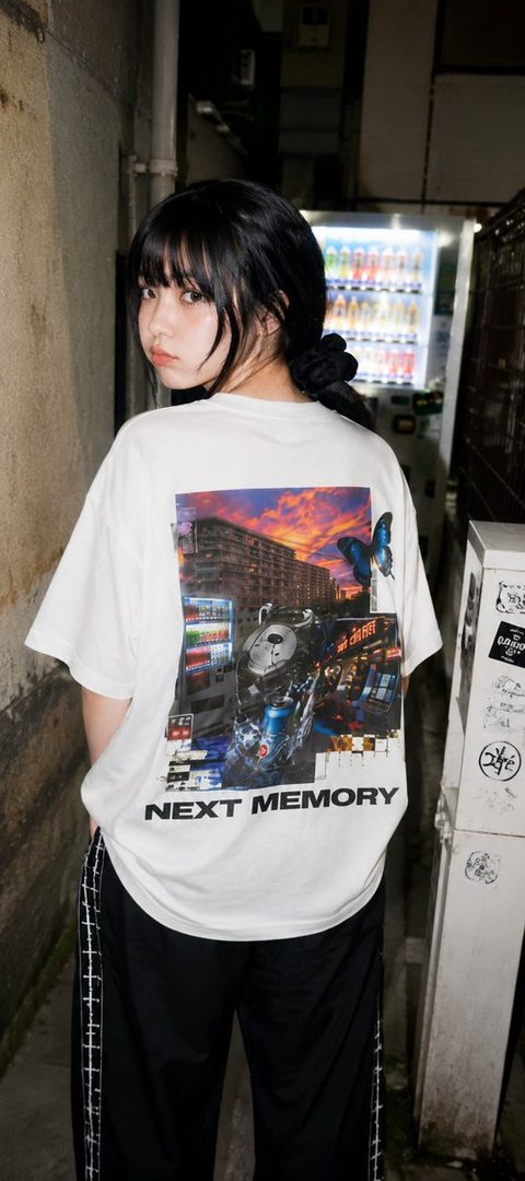 A young woman with black hair and bangs, wearing a white graphic t-shirt and black pants, stands in a narrow alleyway at night with a vending machine in the background.