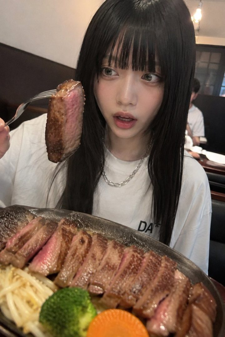 A young woman with long black hair and bangs, wearing a white t-shirt and a silver chain necklace, looks surprised as she holds a fork with a piece of steak.