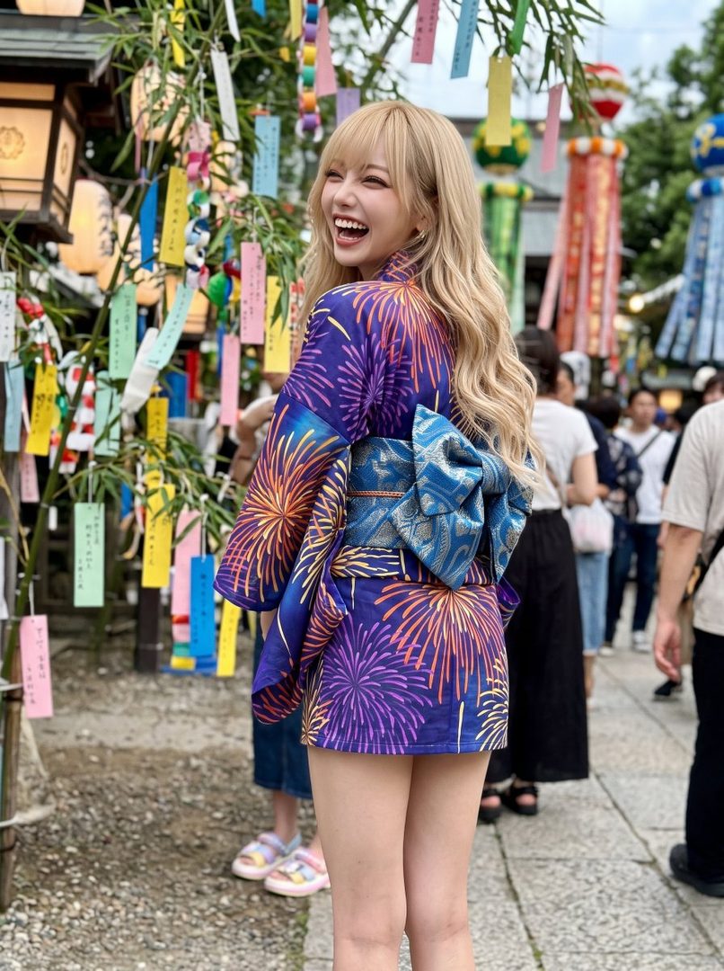 A young Japanese woman with blonde hair and bangs, wearing a purple yukata with a firework pattern and a blue obi, smiles over her shoulder outdoors.