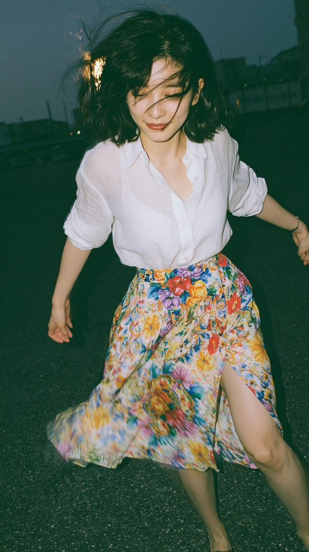 A young Japanese woman with short black hair, wearing a white shirt and a colorful floral skirt, walks outdoors at night.